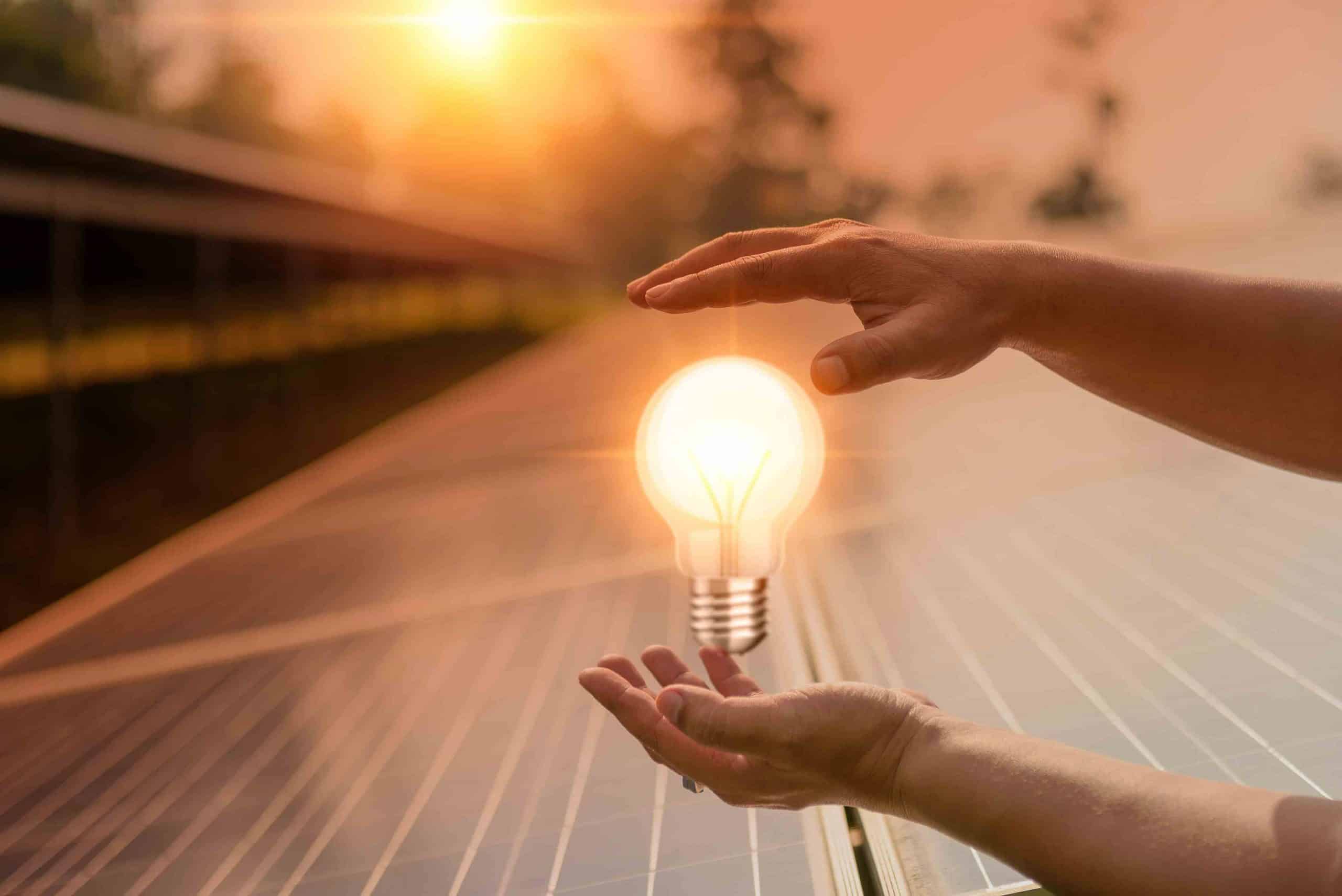Hände halten leuchtende Glühbirne über Solarpaneelen im Sonnenuntergang.