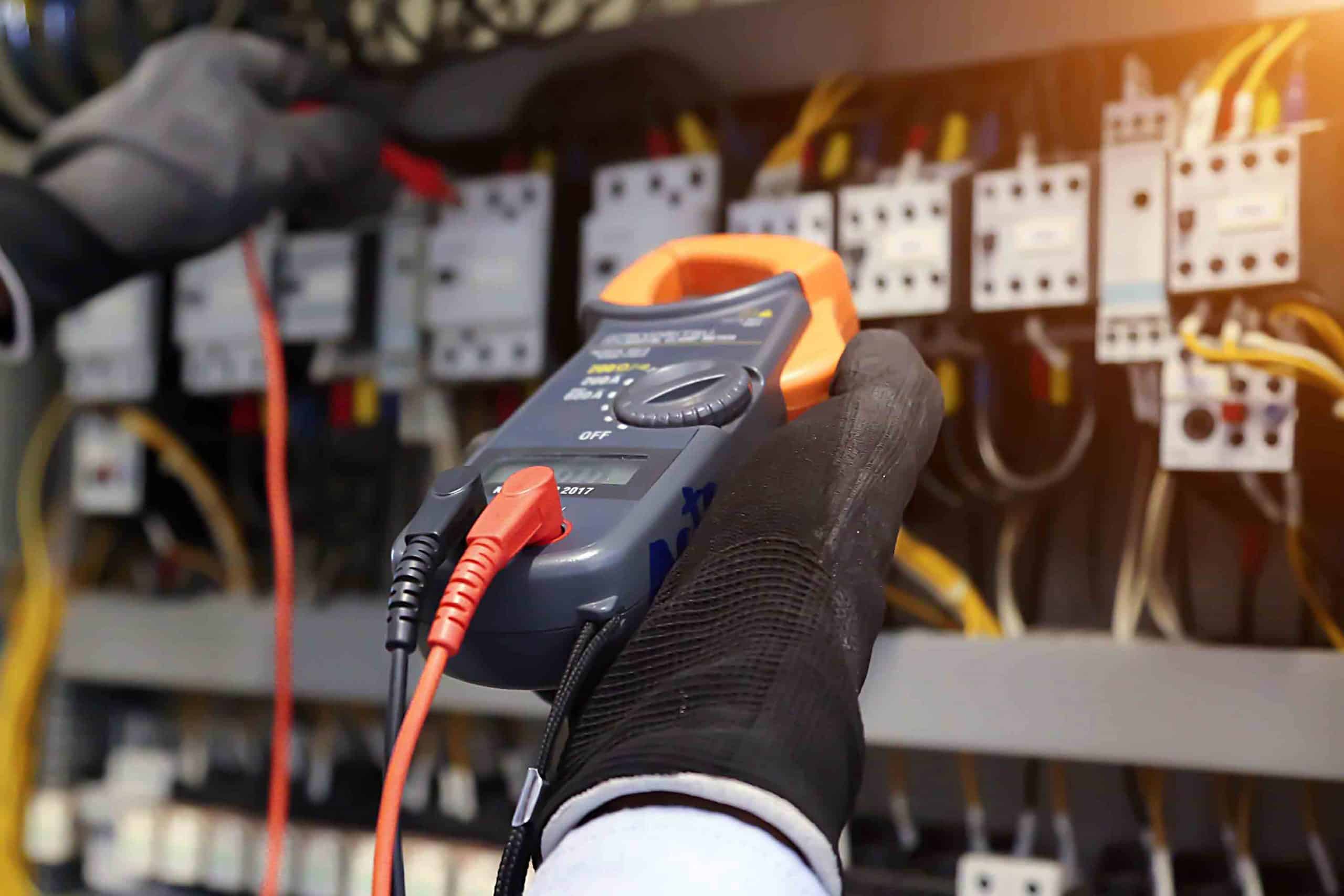 Hand mit Schutzhandschuh hält Multimeter zur Messung an elektrischem Schaltschrank.