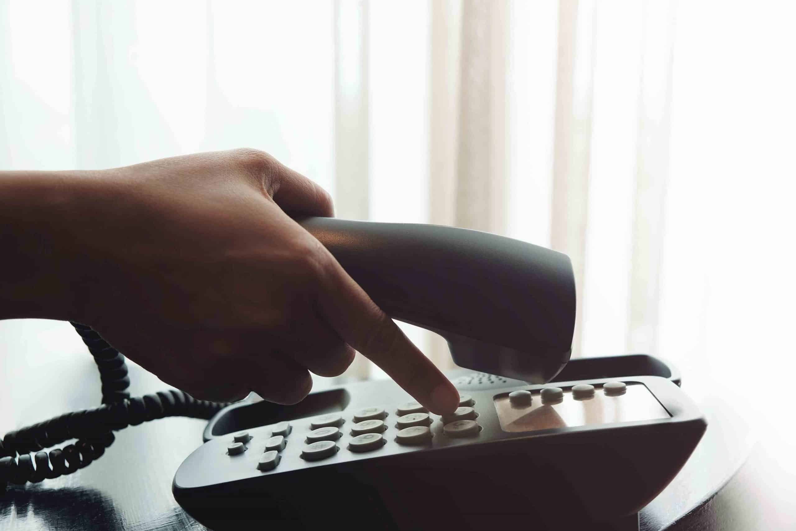 Hand hält Telefonhörer und wählt Nummer auf dem Tastenfeld eines Festnetztelefons.