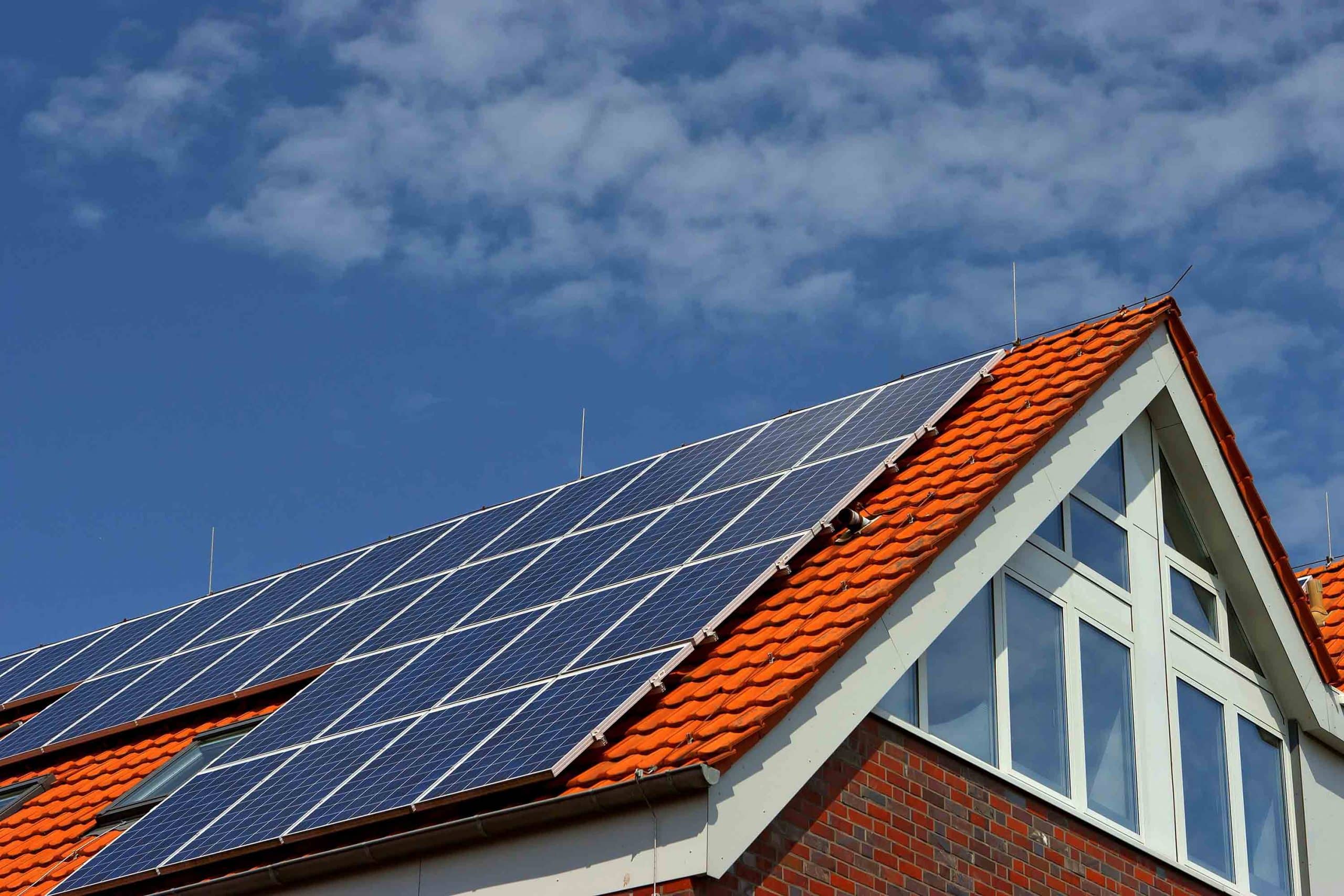 Hausdach mit roten Ziegeln und installierten Solarmodulen bei blauem Himmel mit Wolken.