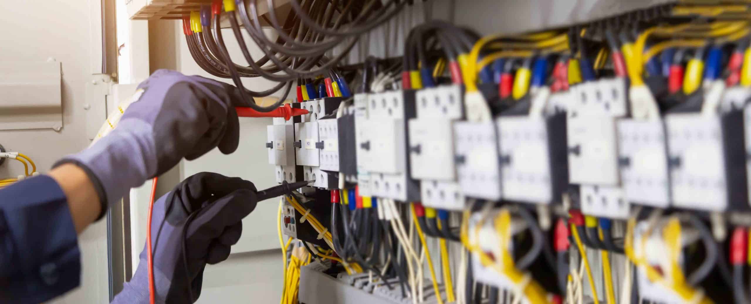 Person mit Schutzhandschuhen misst elektrische Werte an Schalttafel mit Kabeln und Anschlüssen.