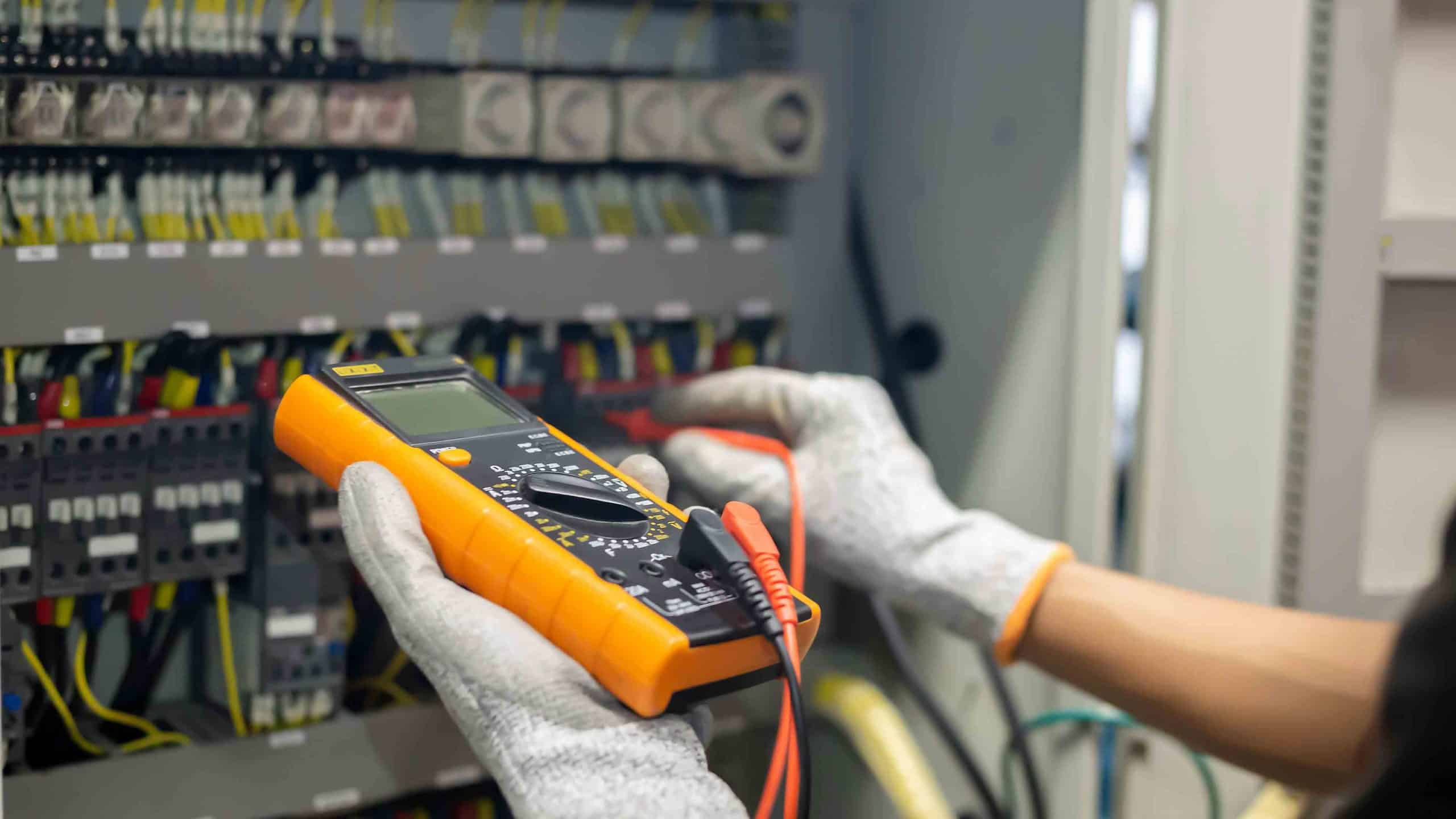 Person mit Schutzhandschuhen misst elektrische Werte an einem Schaltschrank mit Multimeter.